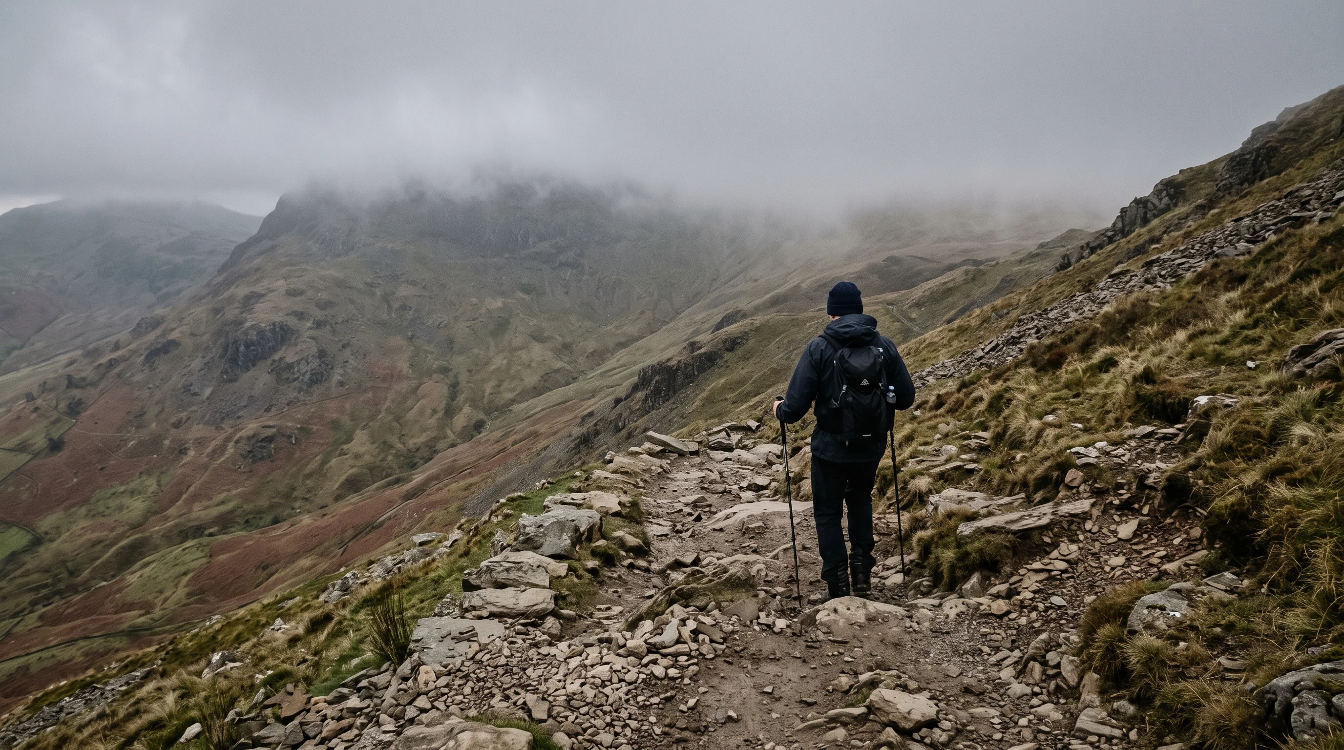 A walker seen from behind, descending a steep, rough stony path off a South Lakes fell under low cloud