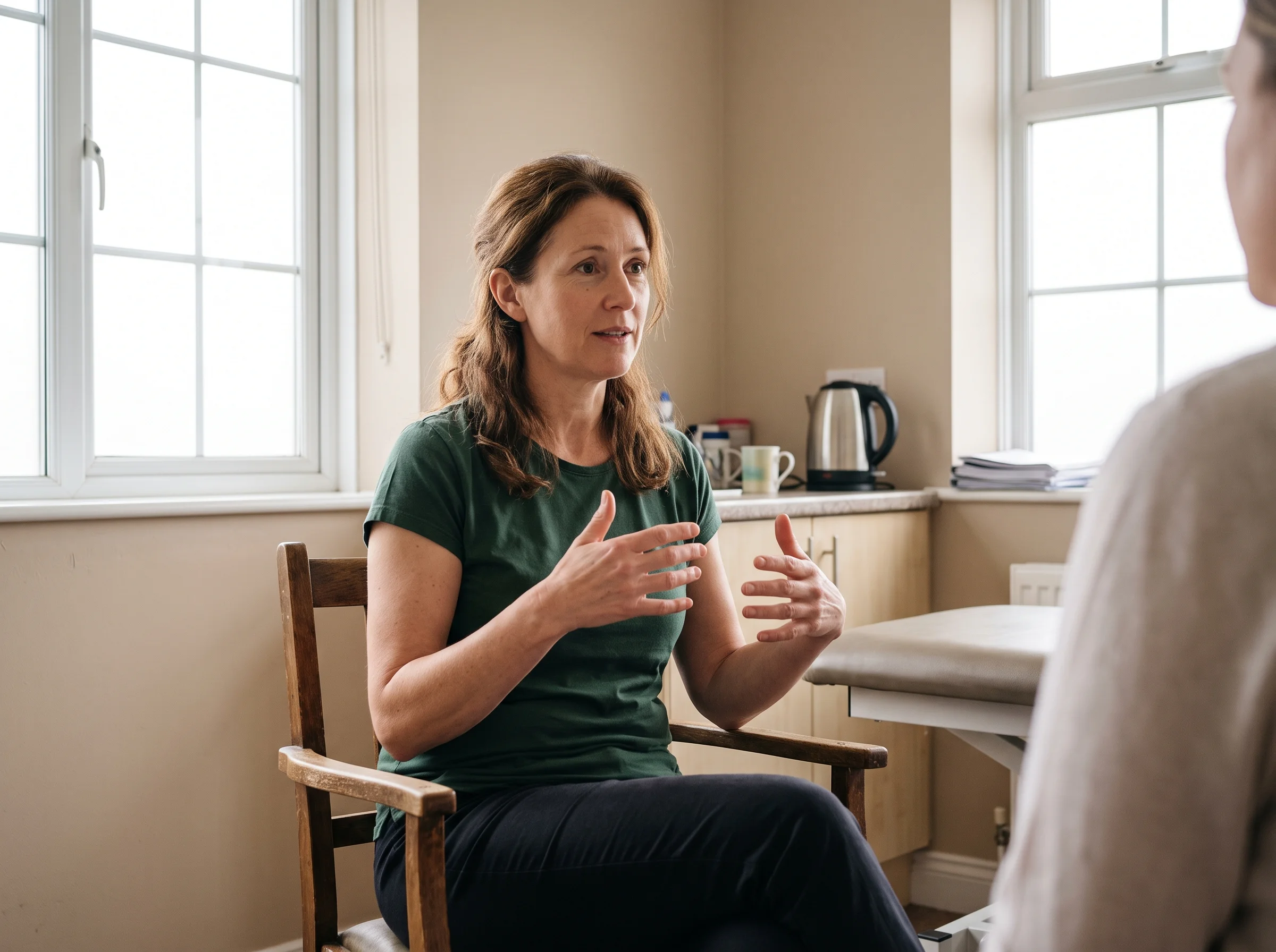 Jo sitting in her ground-floor treatment room, explaining something with her hands, with the kettle on the side table behind her